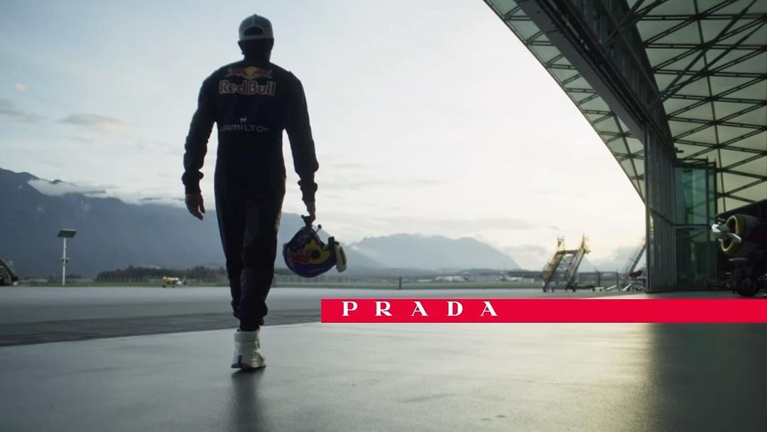 Racer in dark suit walks with helmet on tarmac near hangar with mountains in background, promoting the brand Prada in horizontal red bar.
