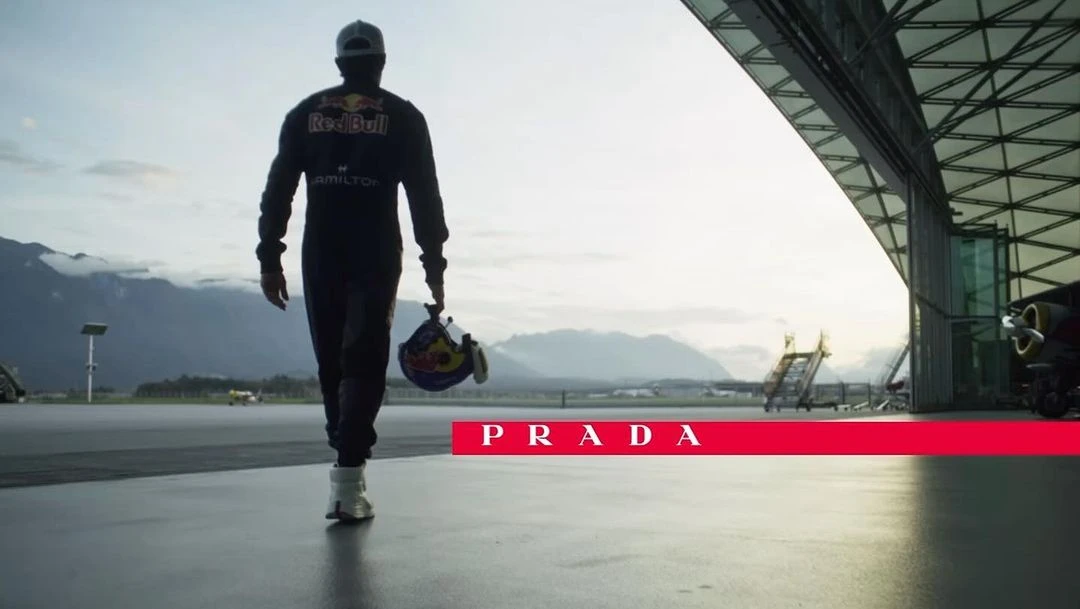 Racer in dark suit walks with helmet on tarmac near hangar with mountains in background, promoting the brand Prada in horizontal red bar.