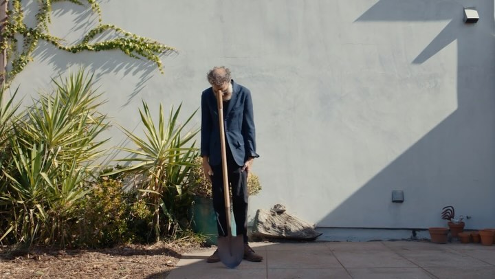 A man in a suit stands in a yard next to a spade, posing outdoors against a light gray wall, with plants and shadows visible.