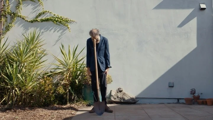 A man in a suit stands in a yard next to a spade, posing outdoors against a light gray wall, with plants and shadows visible.