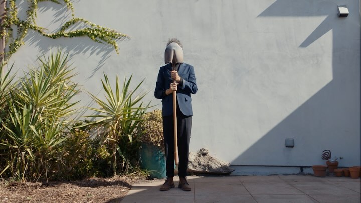 A person in a suit stands outdoors with a shovel covering their face, plants, and a light gray wall in the background.