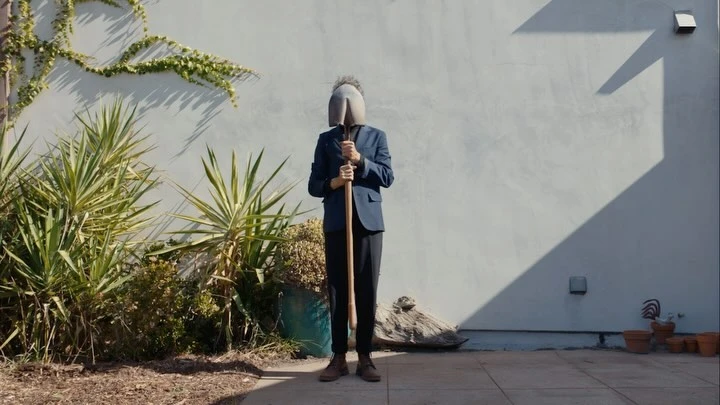 A person in a suit stands outdoors with a shovel covering their face, plants, and a light gray wall in the background.