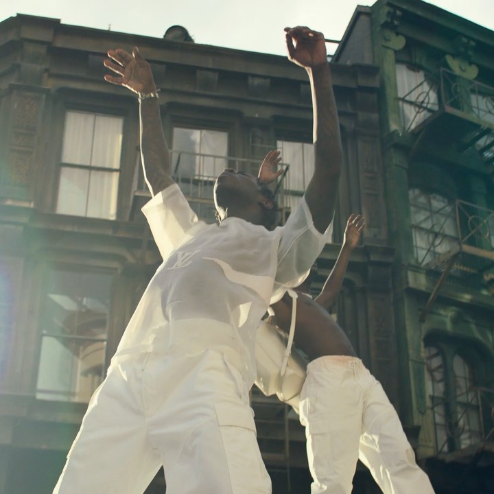 Two adult humans in white outfits pose with arms raised against a backdrop of a city building and sky during daytime.