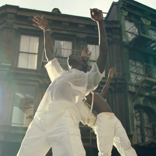 Two adult humans in white outfits pose with arms raised against a backdrop of a city building and sky during daytime.