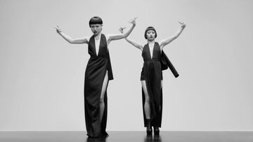 Two women in long black dresses strike a pose on a gray background. One has a jacket draped over her shoulder in a studio shot.