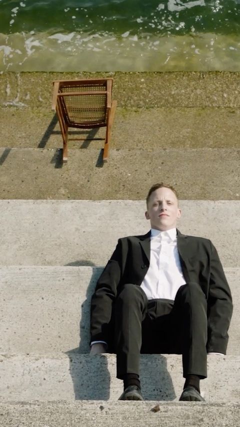 Man in a black suit sits on concrete steps near a body of water with a wicker chair nearby, captured in natural light.