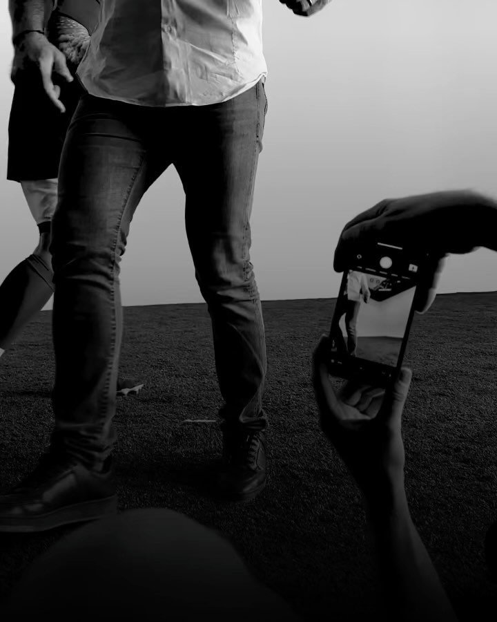 Black and white photo of a man in jeans and a white shirt standing on grass, captured by a person holding a phone.