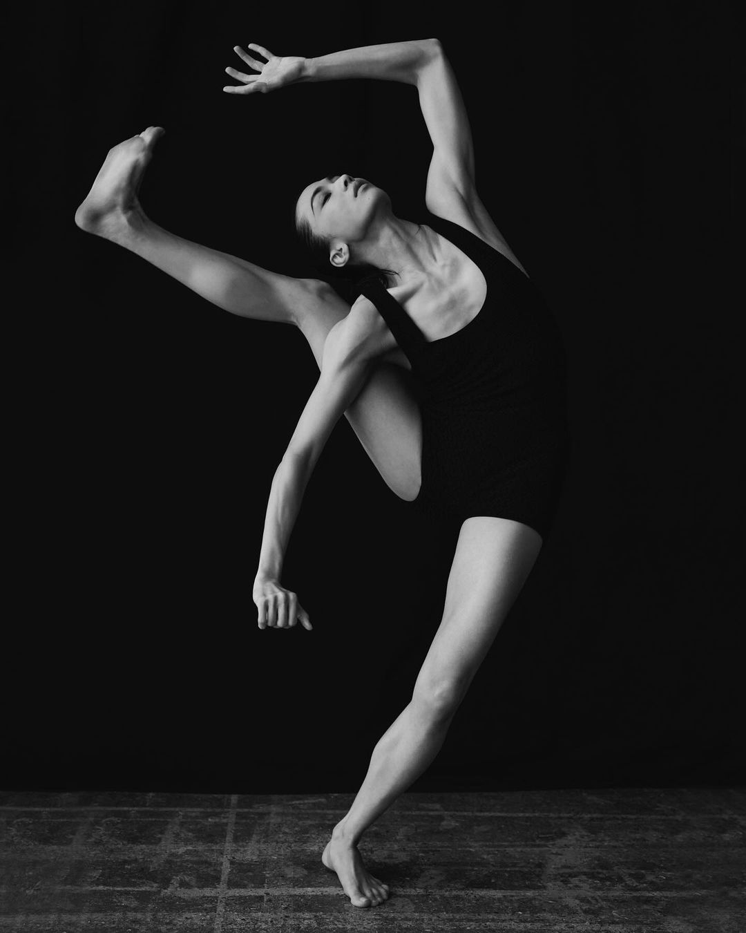 A woman in a black bodysuit strikes an impressive dance pose in a monochrome studio photograph, showcasing her flexibility and strength.