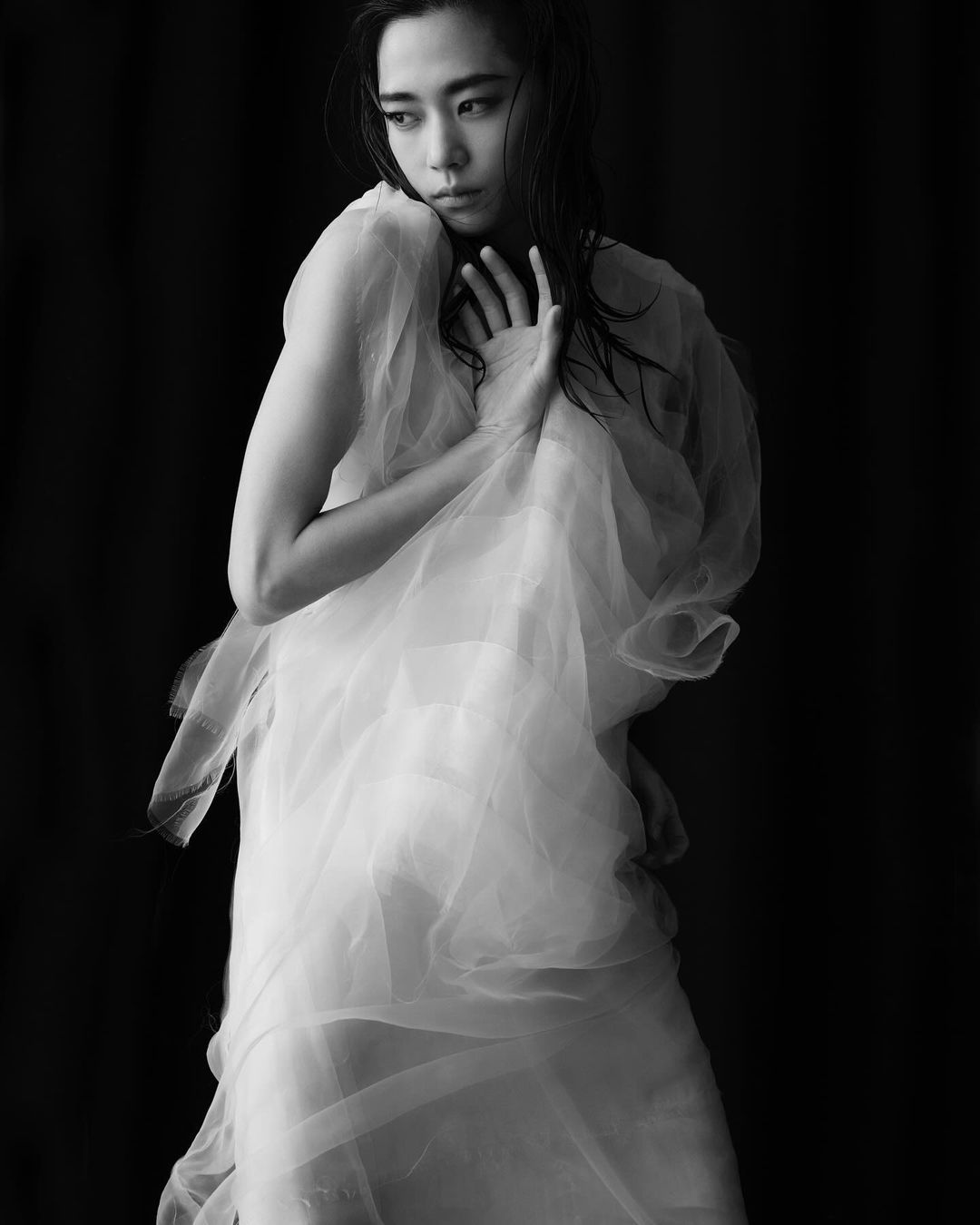 Black-and-white portrait of an Asian woman wrapped in a white sheer fabric, gazing off-camera, in a studio setting with dark background.