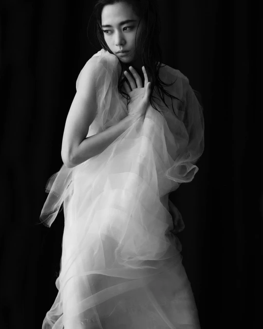 Black-and-white portrait of an Asian woman wrapped in a white sheer fabric, gazing off-camera, in a studio setting with dark background.