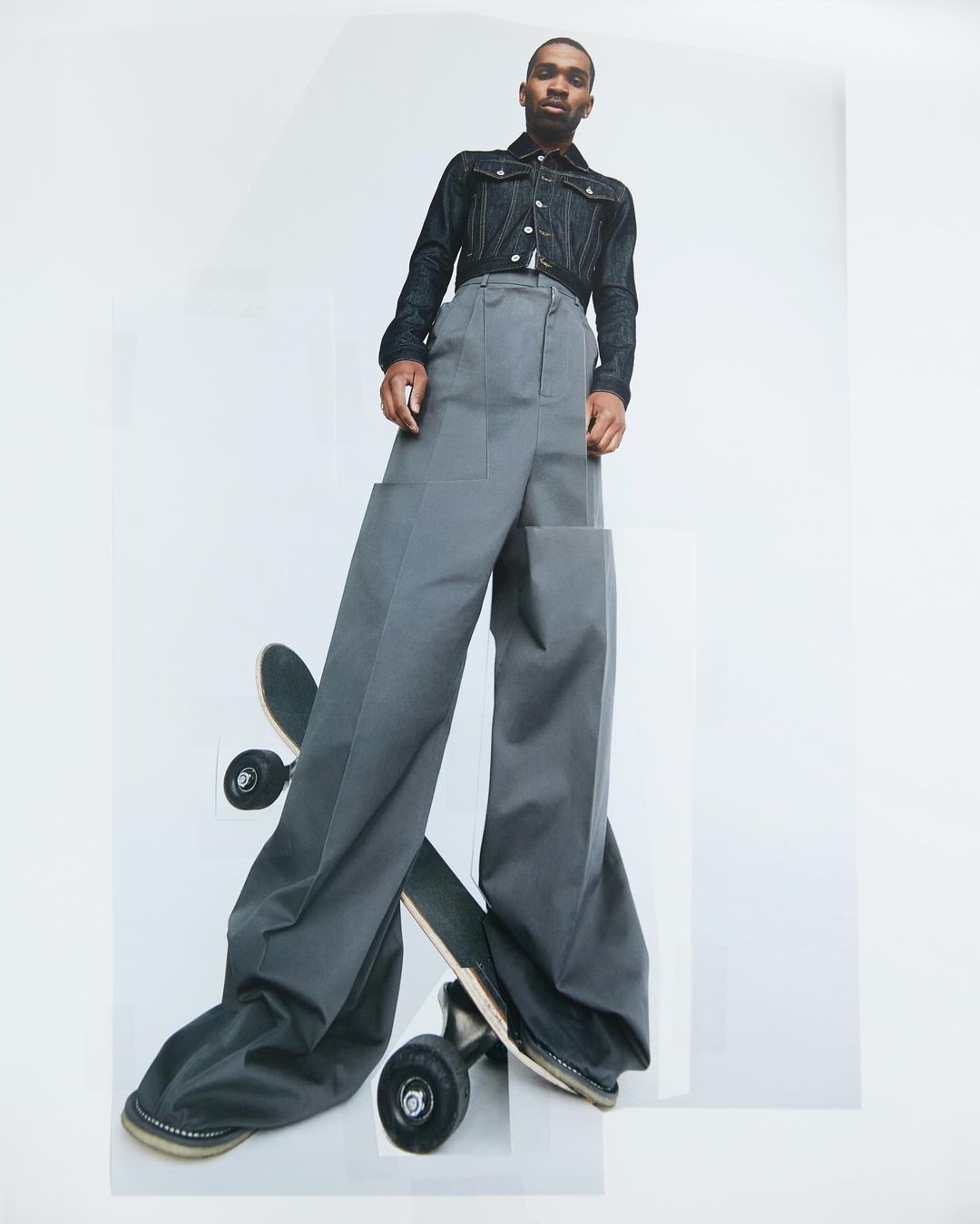A young Black man in denim jacket and oversized grey trousers stands above a skateboard in a studio shot against a bright white background.