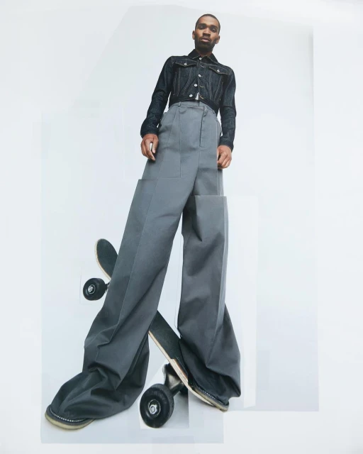 A young Black man in denim jacket and oversized grey trousers stands above a skateboard in a studio shot against a bright white background.