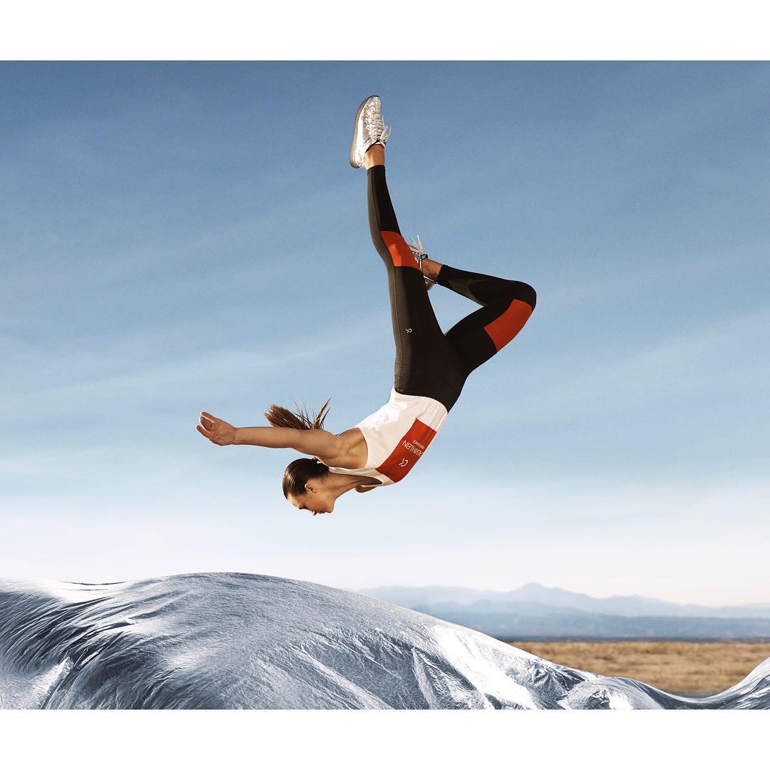 A woman in athletic wear is captured mid-air, performing a flip against a blue sky with mountains and a white tarp in the background.