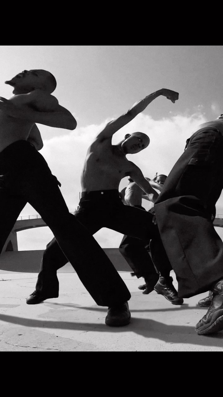 Group of bare-chested men in black pants dancing outdoors under bright sky, captured in black and white photography.