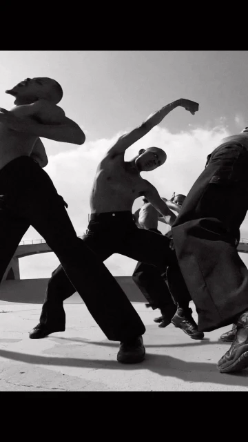Group of bare-chested men in black pants dancing outdoors under bright sky, captured in black and white photography.
