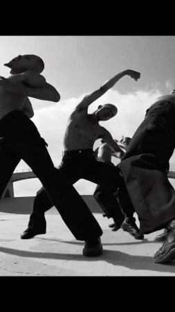 Group of bare-chested men in black pants dancing outdoors under bright sky, captured in black and white photography.