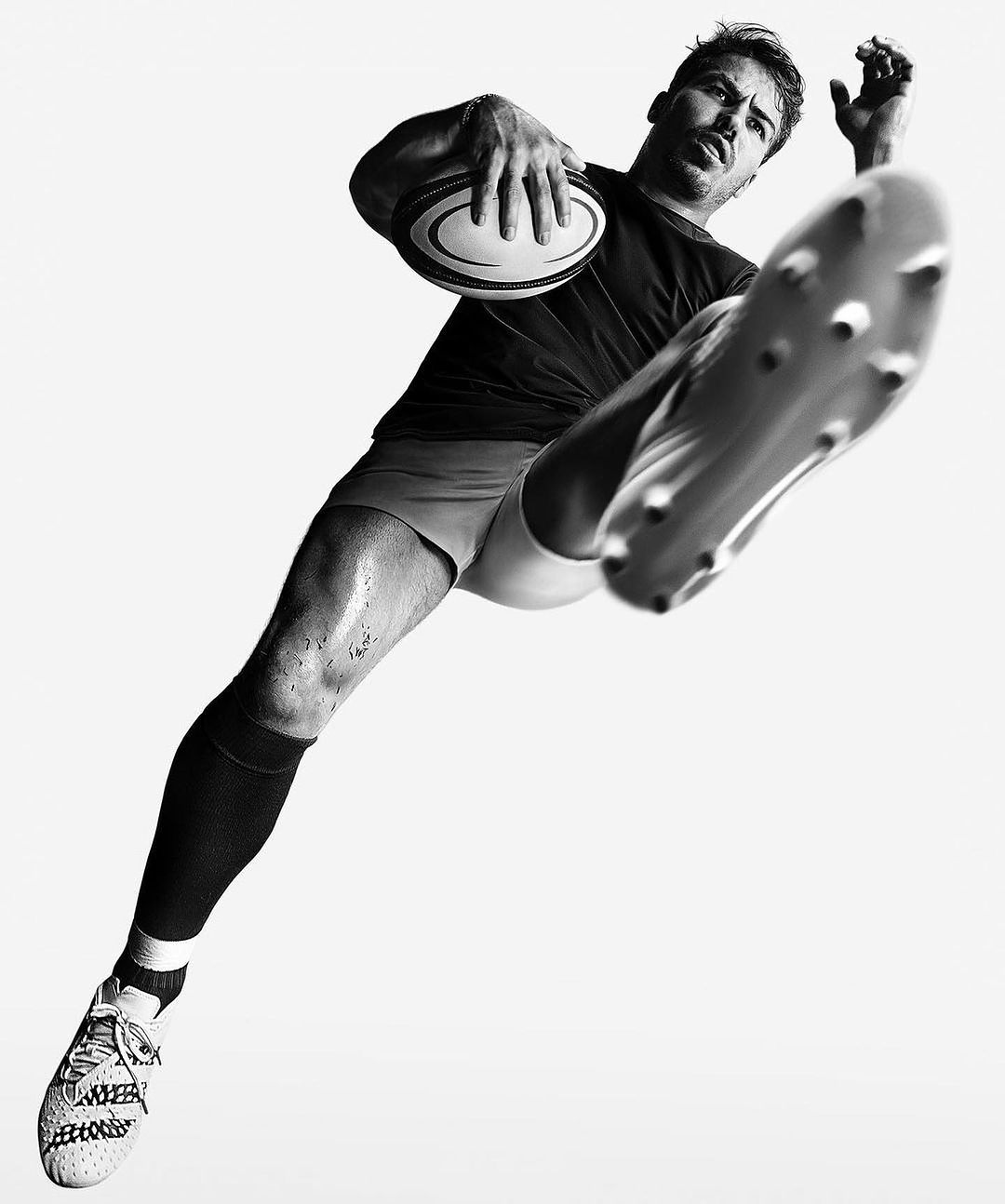 Monochromatic image of a rugby player in action, with a dynamic pose, holding a rugby ball, and wearing sports gear against a white background.