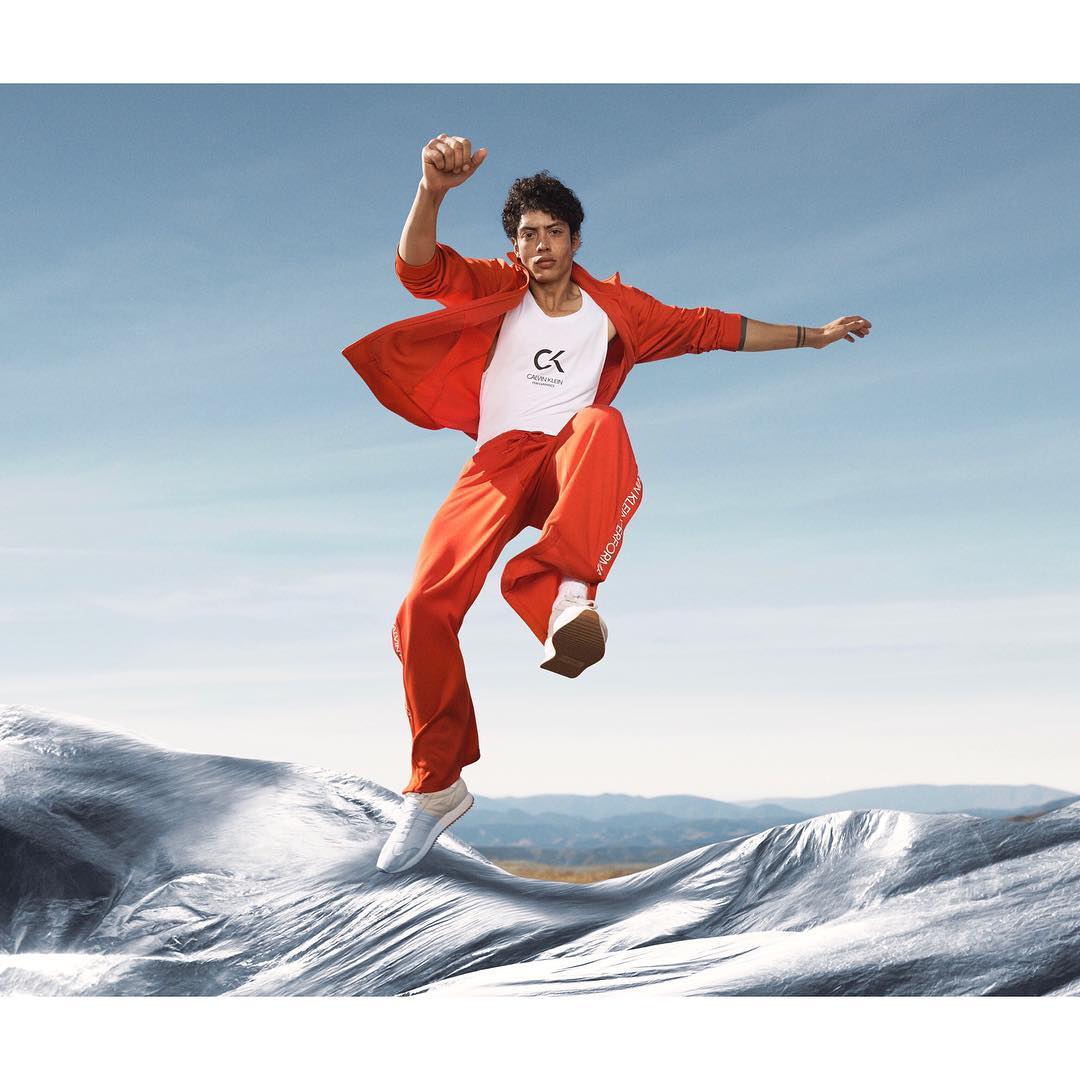 Man in motion wearing a red jacket and pants with white sneakers, leaping over a reflective surface under a blue sky.