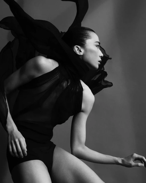 Black and white portrait of an Asian female model in artistic pose, draped with flowing black fabric, side profile in studio setting.