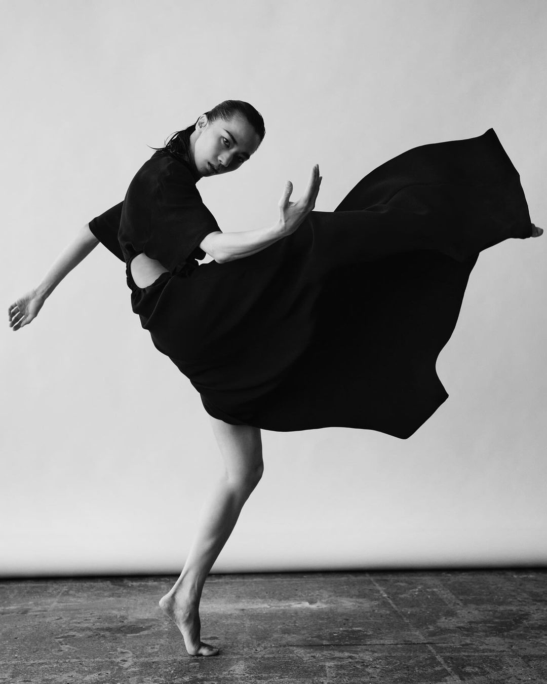 Monochrome image of a barefoot woman in dynamic pose, wearing a black dress with open back, against a neutral backdrop. Fashion and movement are key.