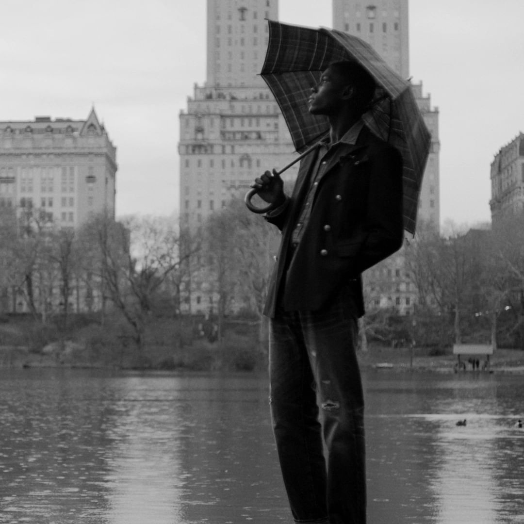 Black-and-white shot of a Black man holding an umbrella in front of a lake and urban buildings on a cloudy day. He looks up.