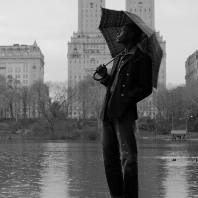 Black-and-white shot of a Black man holding an umbrella in front of a lake and urban buildings on a cloudy day. He looks up.