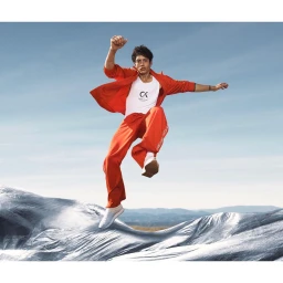 A man wearing an orange tracksuit and white tank top jumps mid-air against a clear blue sky and a textured silver backdrop.