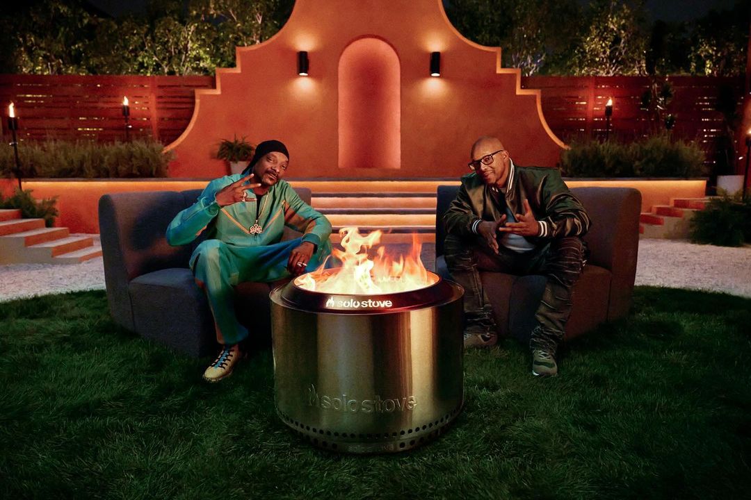 Snoop Dogg and Warren G sit by a lit Solo Stove fire pit on a grassy lawn at night, with outdoor furniture and landscape lighting.
