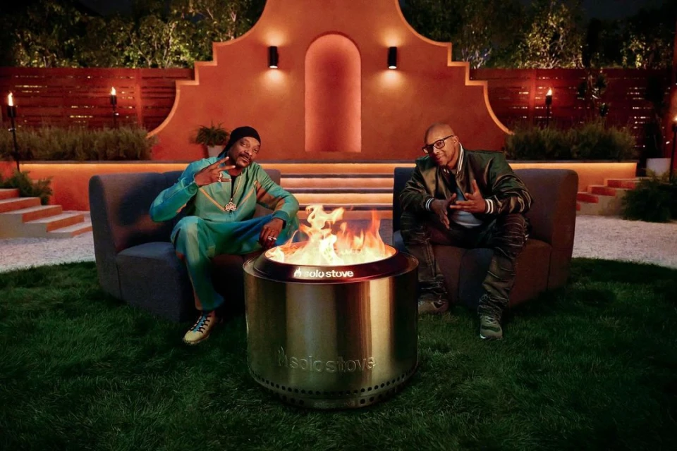 Snoop Dogg and Warren G sit by a lit Solo Stove fire pit on a grassy lawn at night, with outdoor furniture and landscape lighting.