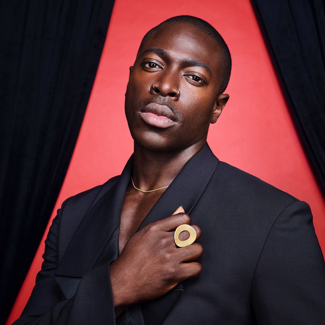 Portrait of a Black man in a black suit against a red background, wearing a gold ring and necklace, looking directly at the camera.