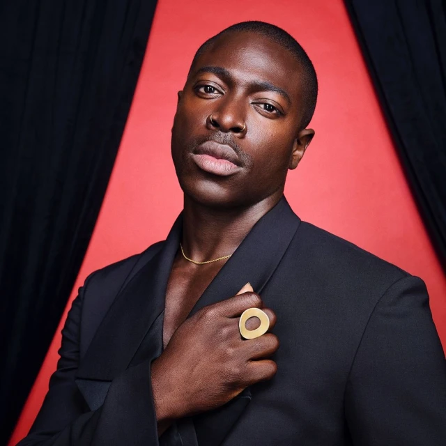 Portrait of a Black man in a black suit against a red background, wearing a gold ring and necklace, looking directly at the camera.