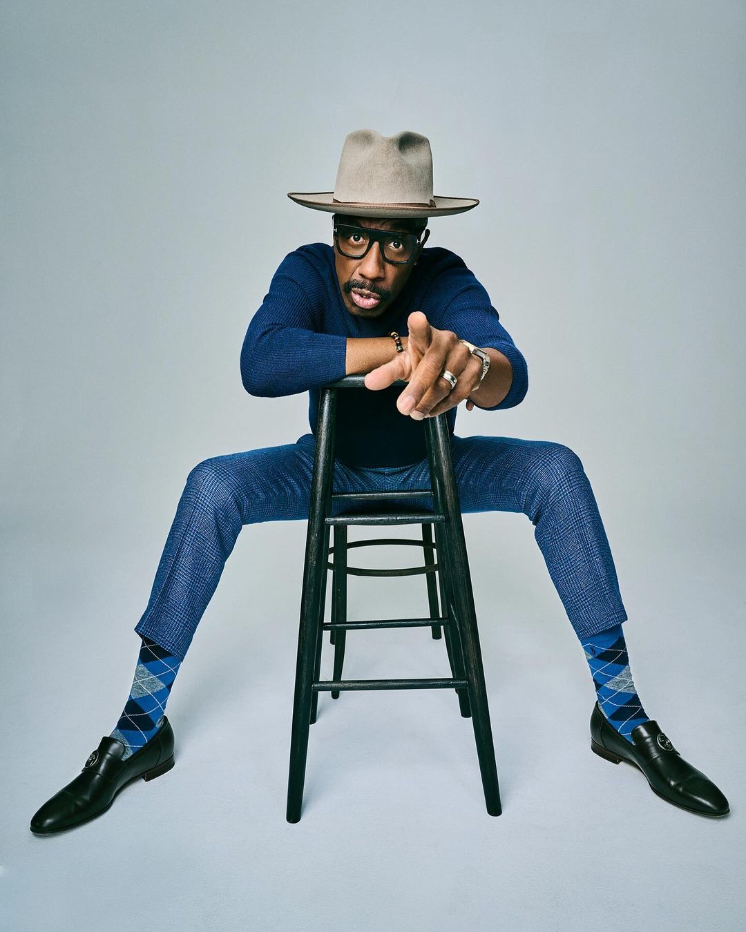 Stylish man in a hat and blue suit sits on a stool, pointing forward in a studio shot against a neutral background, exuding confidence.