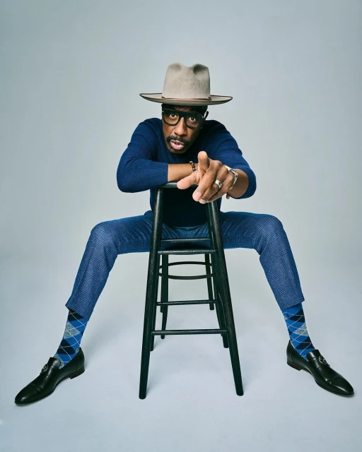 Stylish man in a hat and blue suit sits on a stool, pointing forward in a studio shot against a neutral background, exuding confidence.