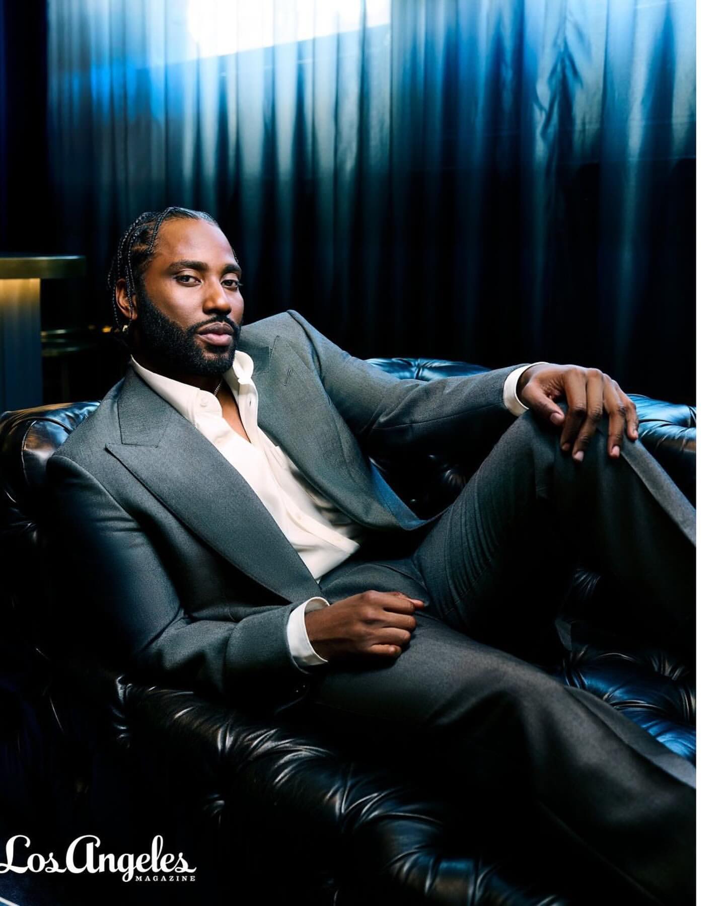 Portrait of a stylish adult man in a grey suit reclining on a black leather sofa, with a dark blue background and diffused lighting.