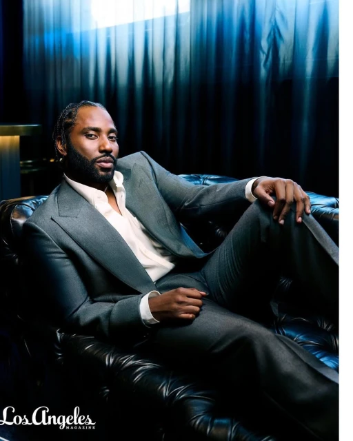 Portrait of a stylish adult man in a grey suit reclining on a black leather sofa, with a dark blue background and diffused lighting.
