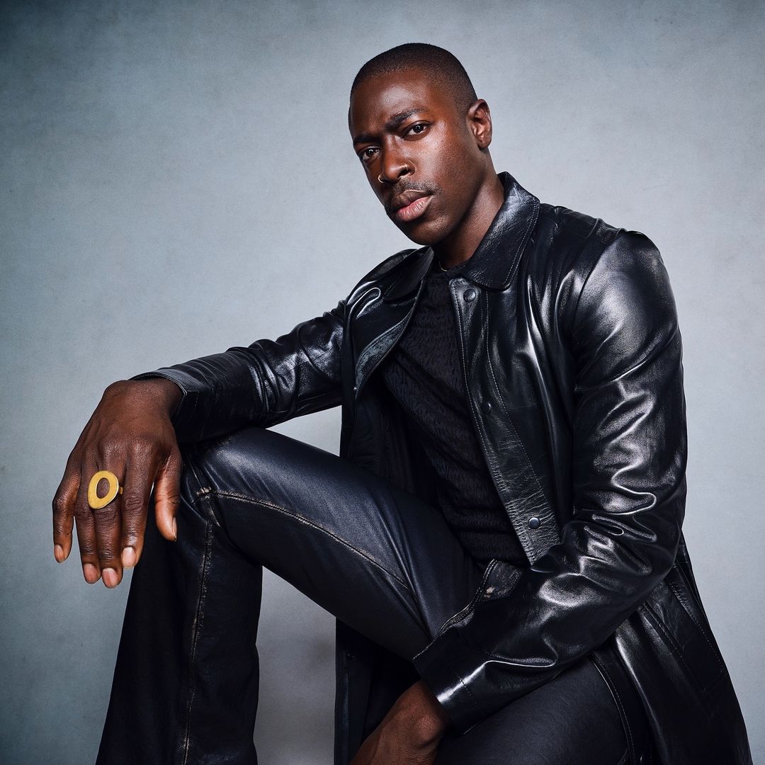 Portrait of a handsome African-American man in a black leather coat and pants, sitting against a grey backdrop and wearing a gold ring.