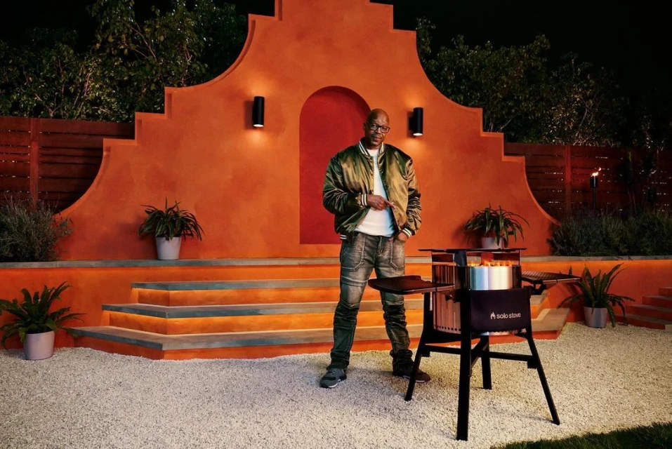 Adult male posing near a fire pit on a gravel patio with outdoor furniture and a warm-toned stucco wall featuring sconce lighting.