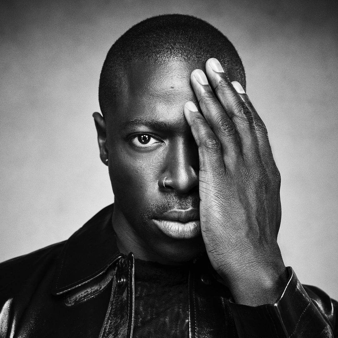 Monochromatic portrait of a Black man with a nose ring, covering half his face with his hand. He wears a leather jacket.