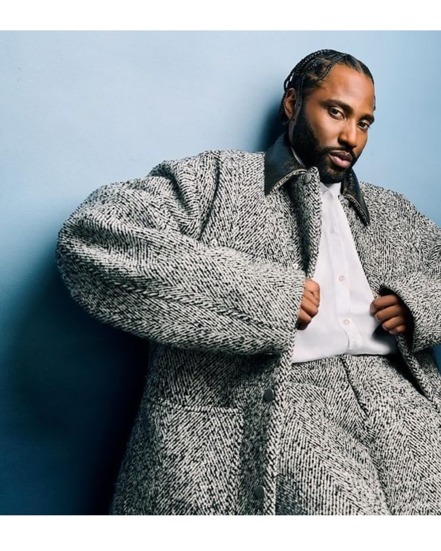 Adult male with braided hair poses in a grey tweed suit, white dress shirt, and black leather collar against a blue-grey backdrop.