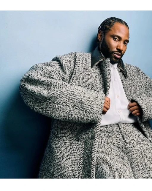 Adult male with braided hair poses in a grey tweed suit, white dress shirt, and black leather collar against a blue-grey backdrop.