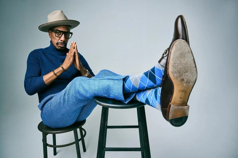 Stylish adult male sits on a stool wearing a hat, blue sweater, and patterned pants, with dress shoes showing soles in a studio shot.