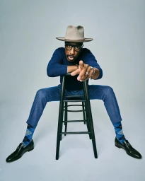 Man in blue sweater and fedora hat sitting backwards on a black stool pointing towards camera against a white backdrop.