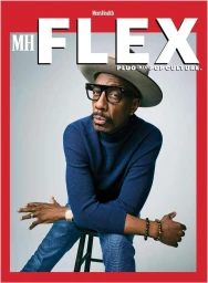 J.B. Smoove wearing a blue turtleneck sweater, fedora, and thick-rimmed glasses seated against a grey backdrop for Men's Health Flex.