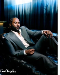 John David Washington seated on a black leather sofa wearing a grey suit and white shirt, photographed by Jai Lennard.