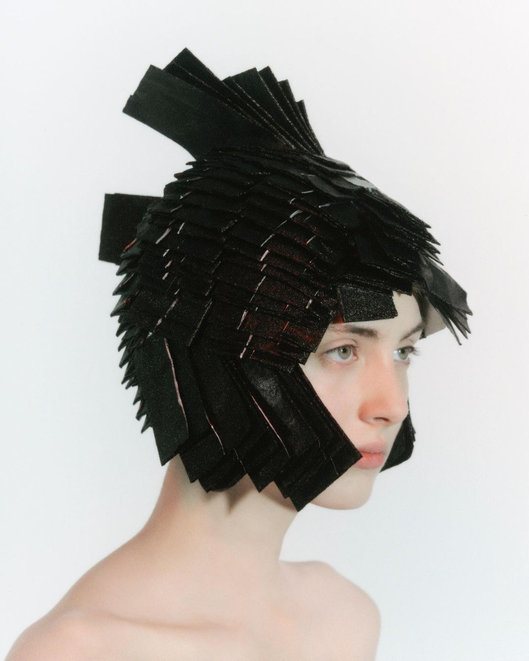 Profile shot of a person with a black geometric headdress on a white background. Unusual headwear fashion editorial portrait.