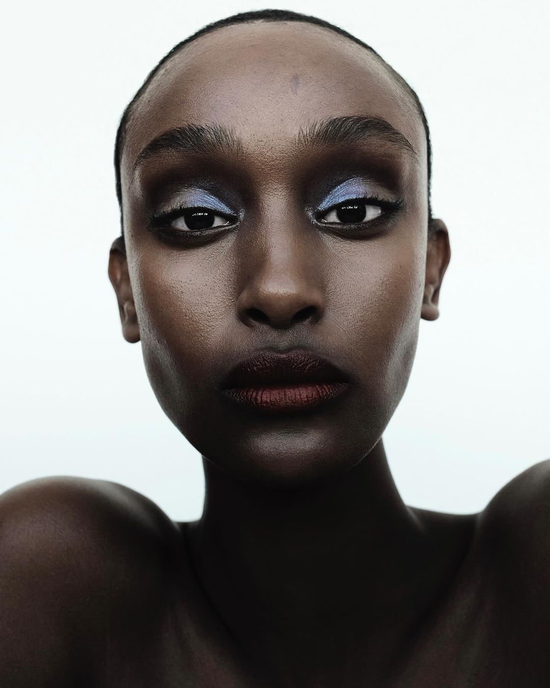 Close-up beauty shot of an adult female with dark skin and blue eye shadow, facing forward against a white background in portrait orientation.
