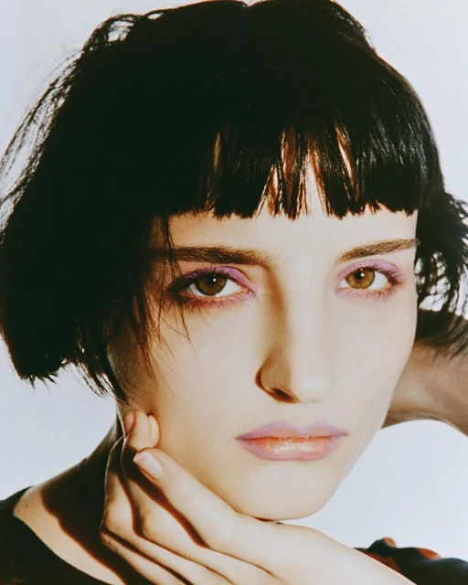 Close-up portrait of a woman with short black hair and light skin, wearing makeup. She is holding her hand to her face.