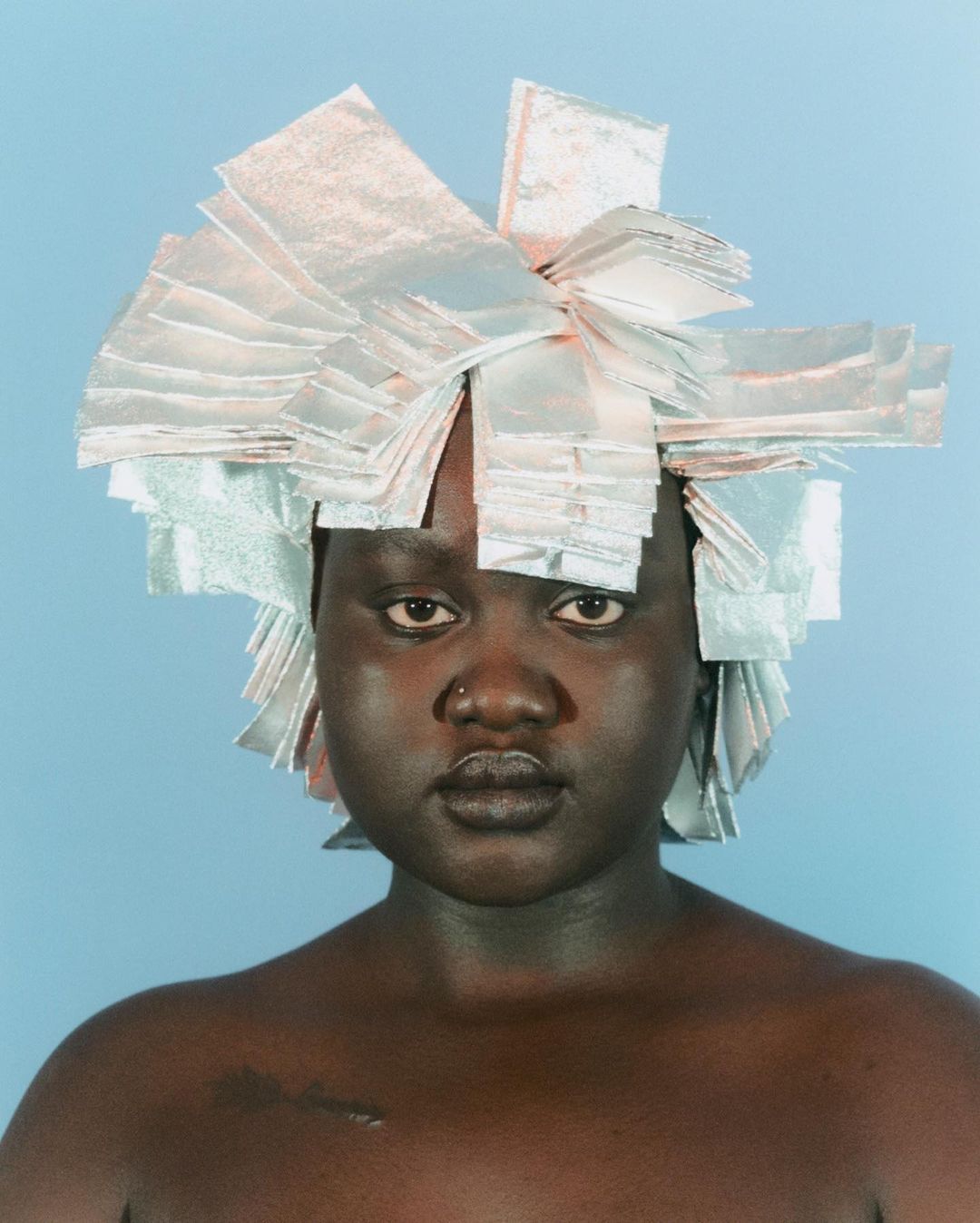 Striking portrait of a young Black woman with dark skin and short hair adorned with silver foils, set against a soft blue backdrop.