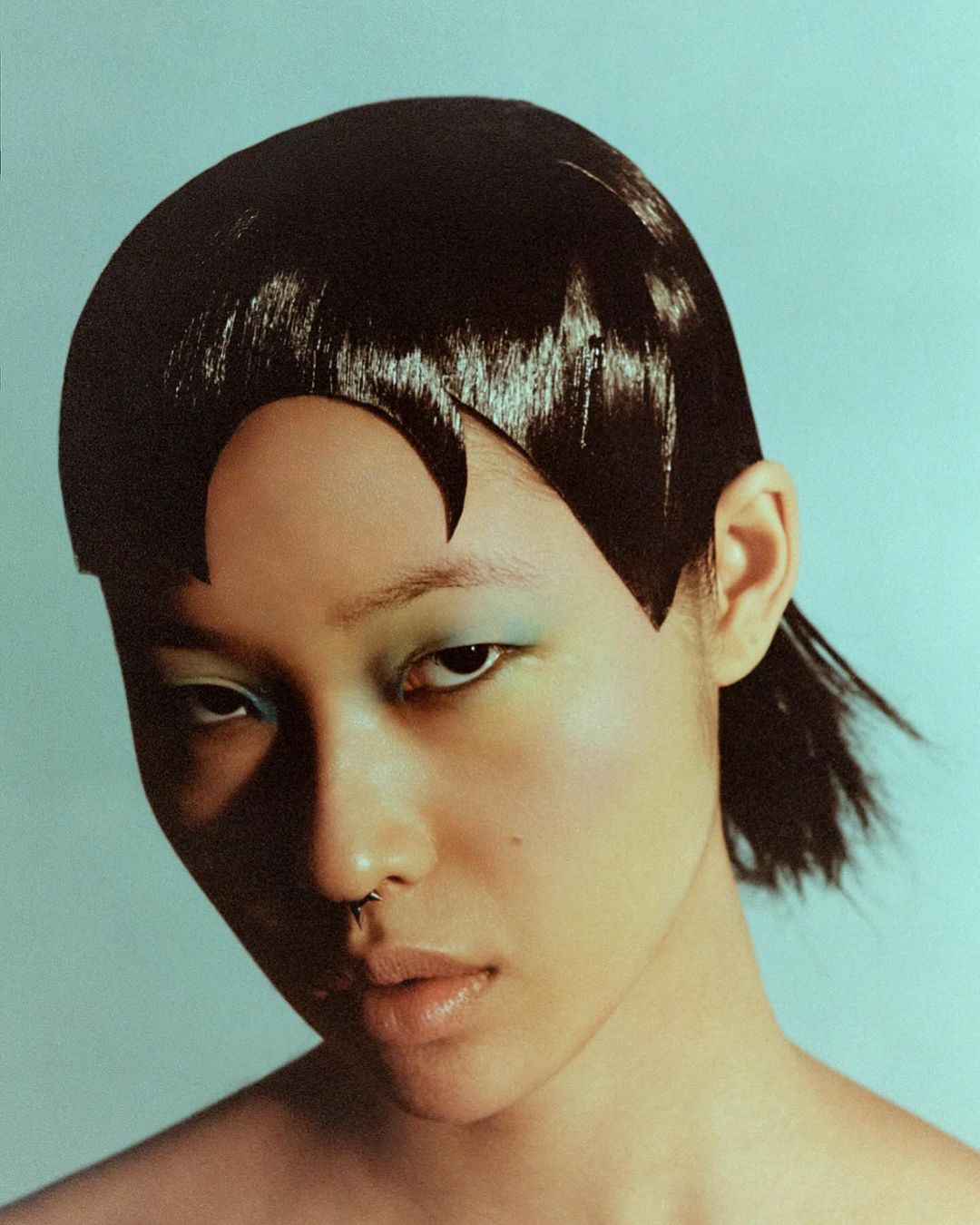 A young Asian woman with dark, stylized hair and green eye makeup, stares intensely at the camera against a pale blue background.