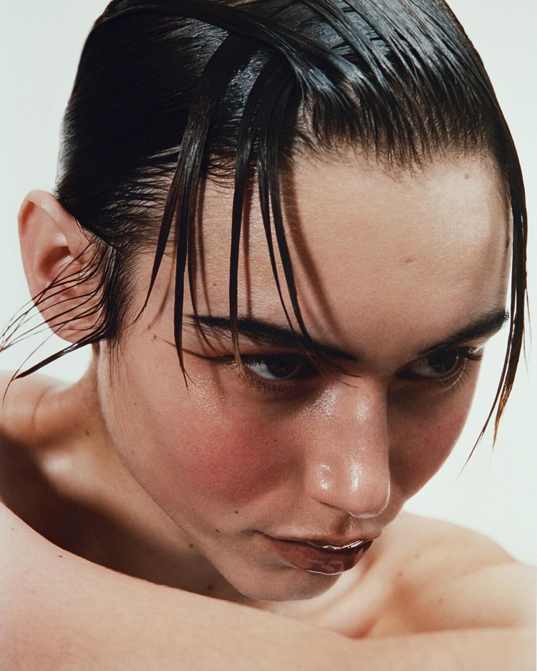 Close-up beauty shot of a person with wet dark hair and glossy lips against a clean white backdrop, showcasing skincare and grooming.